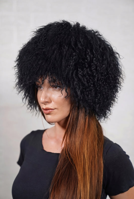 Mongolian Sheepskin Hat Black — Curly Lamb Fur, One Size — three-quarter view worn by a woman, long curly deep jet black Tibetan lamb fur framing the face, TatiFurs