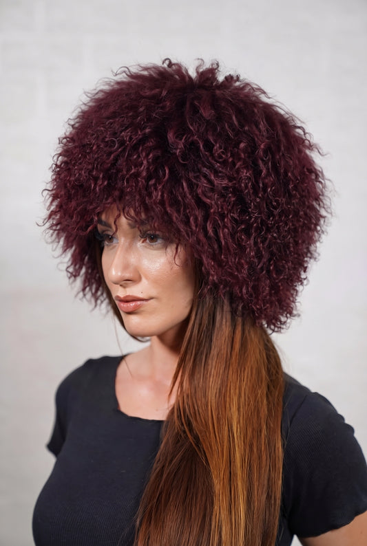 Mongolian Sheepskin Hat Burgundy — Curly Lamb Fur, One Size — three-quarter view worn by a woman, long curly deep oxblood burgundy Tibetan lamb fur framing the face, TatiFurs