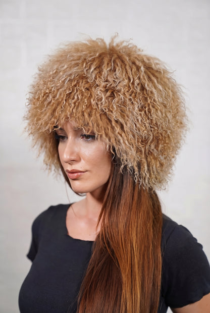 Mongolian Sheepskin Hat Camel Brown — Curly Lamb Fur, One Size — three-quarter view worn by a woman, long curly warm natural camel brown Tibetan lamb fur framing the face, TatiFurs