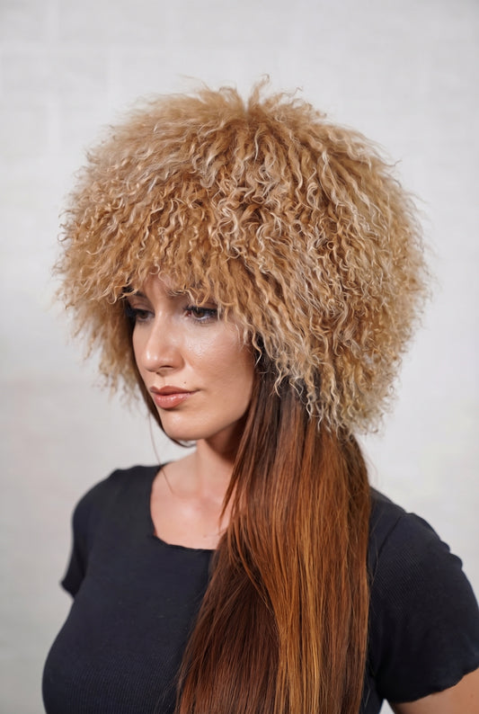 Mongolian Sheepskin Hat Camel Brown — Curly Lamb Fur, One Size — three-quarter view worn by a woman, long curly warm natural camel brown Tibetan lamb fur framing the face, TatiFurs