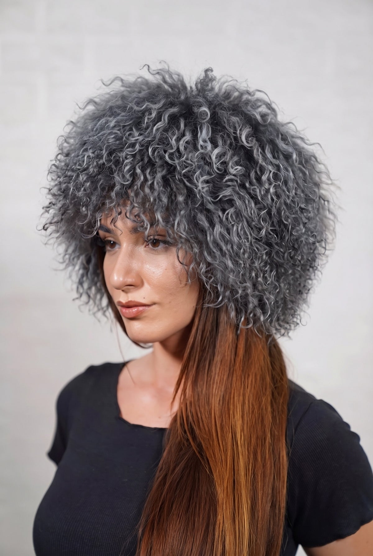 Mongolian Sheepskin Hat Grey — Curly Lamb Fur, One Size — three-quarter view worn by a woman, long curly smoky charcoal grey Tibetan lamb fur framing the face, TatiFurs