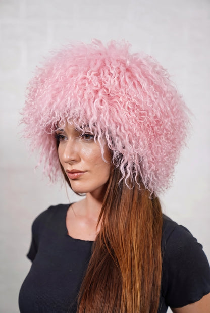 Mongolian Sheepskin Hat Pink — Curly Lamb Fur, One Size — three-quarter view worn by a woman, long curly pale powder pink Tibetan lamb fur framing the face, TatiFurs