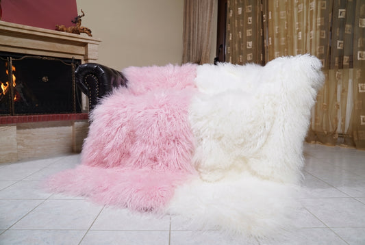 Mongolian Blanket Throw Pink White Two Tone — Wool Sheepskin draped over a dark brown leather Chesterfield sofa beside a lit fireplace — full view of the powder pink and pure white long curly wool, TatiFurs