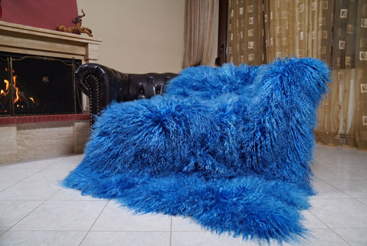 Mongolian Blanket Throw Blue — Wool Sheepskin Full Skin draped over a dark brown leather Chesterfield sofa beside a lit fireplace — full view of the vivid cobalt blue long curly wool, TatiFurs