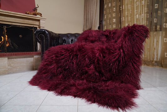 Mongolian Blanket Throw Burgundy — Wool Sheepskin Full Skin draped over a dark brown leather Chesterfield sofa beside a lit fireplace — full view of the deep oxblood burgundy long curly wool, TatiFurs