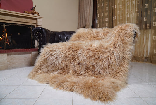 Mongolian Blanket Throw Camel — Wool Sheepskin Full Skin draped over a dark brown leather Chesterfield sofa beside a lit fireplace — full view of the warm golden camel long curly wool, TatiFurs