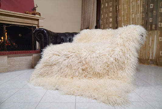 Mongolian Blanket Throw Cream — Wool Sheepskin Full Skin draped over a dark brown leather Chesterfield sofa beside a lit fireplace — full view of the soft ivory cream long curly wool, TatiFurs