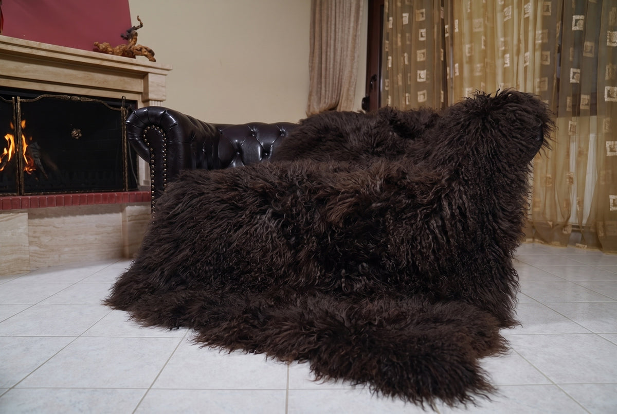 Mongolian Blanket Throw Dark Brown — Wool Sheepskin Full Skin draped over a dark brown leather Chesterfield sofa beside a lit fireplace — full view of the deep espresso dark brown long curly wool, TatiFurs
