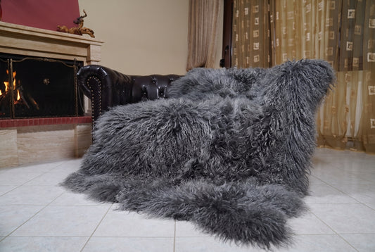 Mongolian Blanket Throw Gray — Wool Sheepskin Full Skin draped over a dark brown leather Chesterfield sofa beside a lit fireplace — full view of the charcoal grey long curly wool, TatiFurs