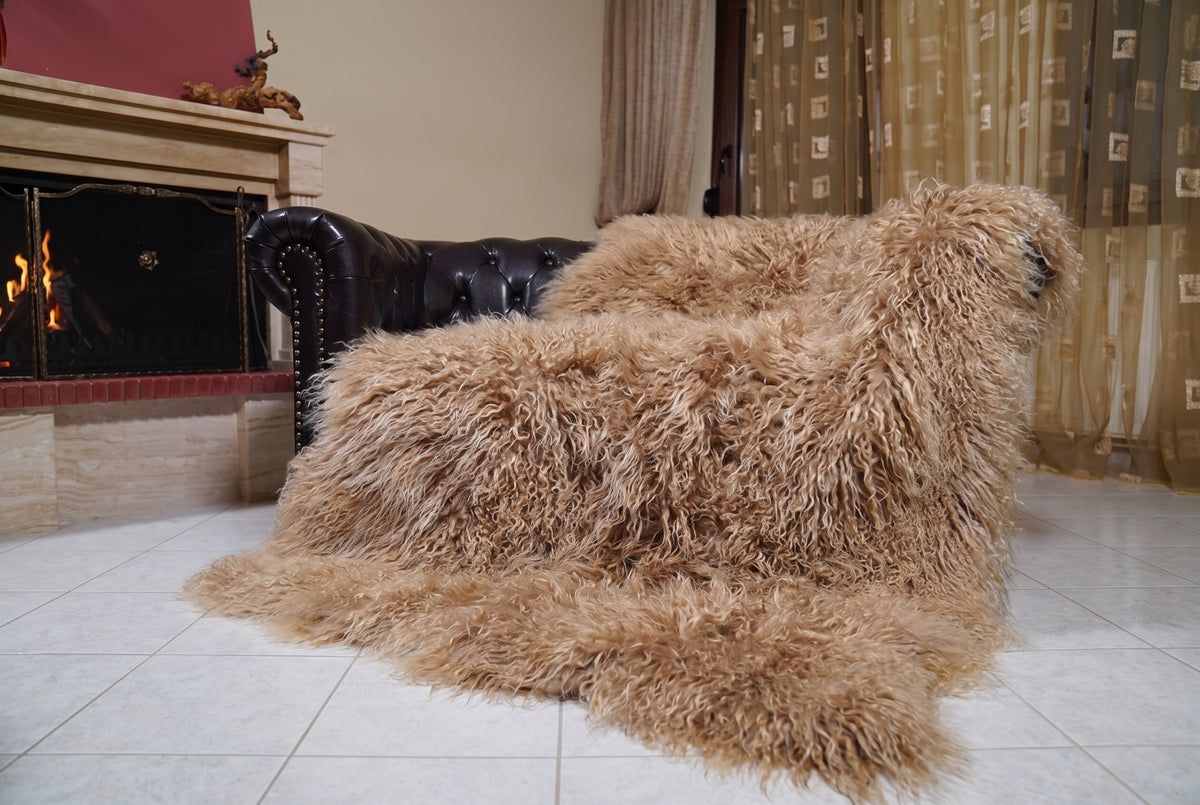 Mongolian Blanket Throw Light Brown — Wool Sheepskin Full Skin draped over a dark brown leather Chesterfield sofa beside a lit fireplace — full view of the milky caramel light brown long curly wool, TatiFurs
