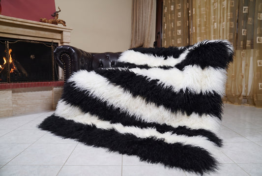 Mongolian Blanket Throw Black White Striped — Wool Sheepskin draped over a dark brown leather Chesterfield sofa beside a lit fireplace — full view of the jet black and pure white long curly wool, TatiFurs