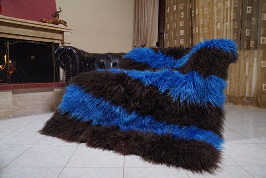 Mongolian Blanket Throw Blue Dark Brown Striped — Wool Sheepskin draped over a dark brown leather Chesterfield sofa beside a lit fireplace — full view of the cobalt blue and espresso brown long curly wool, TatiFurs