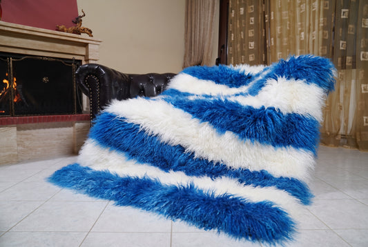 Mongolian Blanket Throw Blue White Striped — Wool Sheepskin draped over a dark brown leather Chesterfield sofa beside a lit fireplace — full view of the cobalt blue and pure white long curly wool, TatiFurs