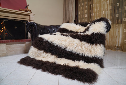 Mongolian Blanket Throw Dark Brown Cream Beige Striped draped over a dark brown leather Chesterfield sofa beside a lit fireplace — full view of the espresso brown and cream beige long curly wool, TatiFurs