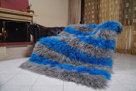 Mongolian Blanket Throw Grey Blue Striped — Wool Sheepskin draped over a dark brown leather Chesterfield sofa beside a lit fireplace — full view of the charcoal grey and cobalt blue long curly wool, TatiFurs