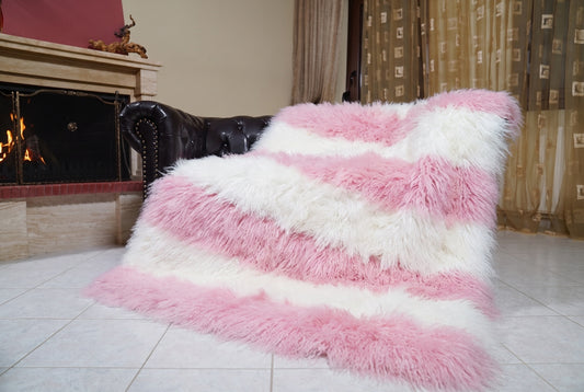 Mongolian Blanket Throw Pink White Striped — Wool Sheepskin draped over a dark brown leather Chesterfield sofa beside a lit fireplace — full view of the powder pink and pure white long curly wool, TatiFurs