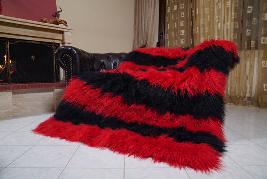 Mongolian Blanket Throw Red Black Striped — Wool Sheepskin draped over a dark brown leather Chesterfield sofa beside a lit fireplace — full view of the scarlet red and jet black long curly wool, TatiFurs