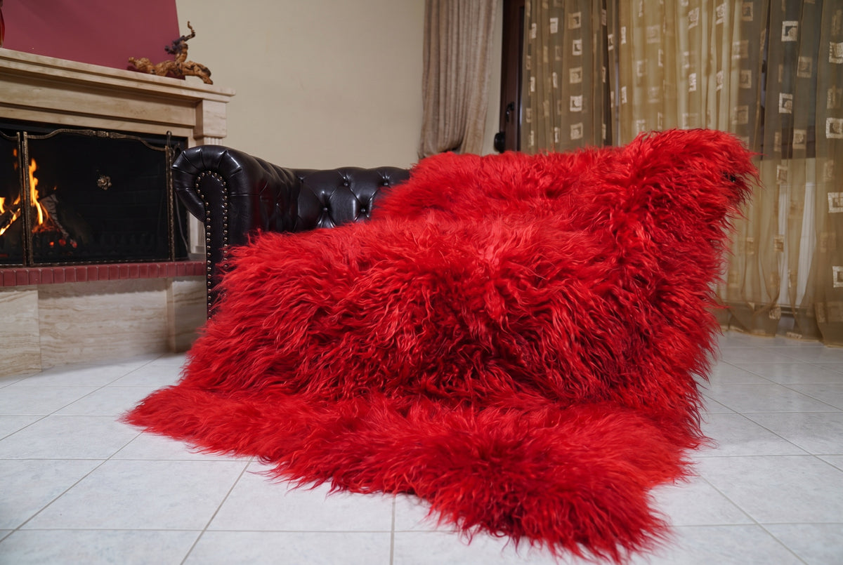 Mongolian Blanket Throw Red — Wool Sheepskin Full Skin draped over a dark brown leather Chesterfield sofa beside a lit fireplace — full view of the vivid scarlet red long curly wool, TatiFurs