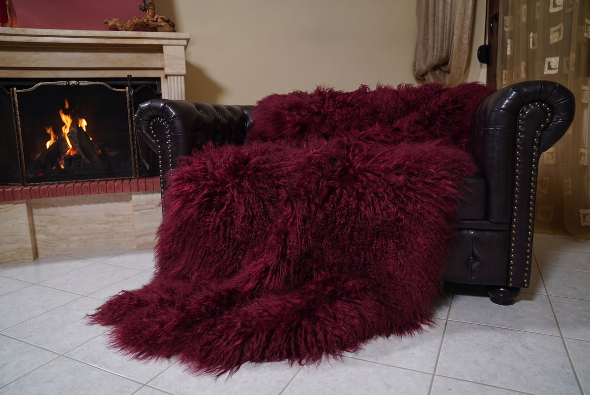 Mongolian Blanket Throw Burgundy — Wool Sheepskin Full Skin angled view on the Chesterfield sofa with the fire glowing behind — deep oxblood burgundy full skin sheepskin, TatiFurs