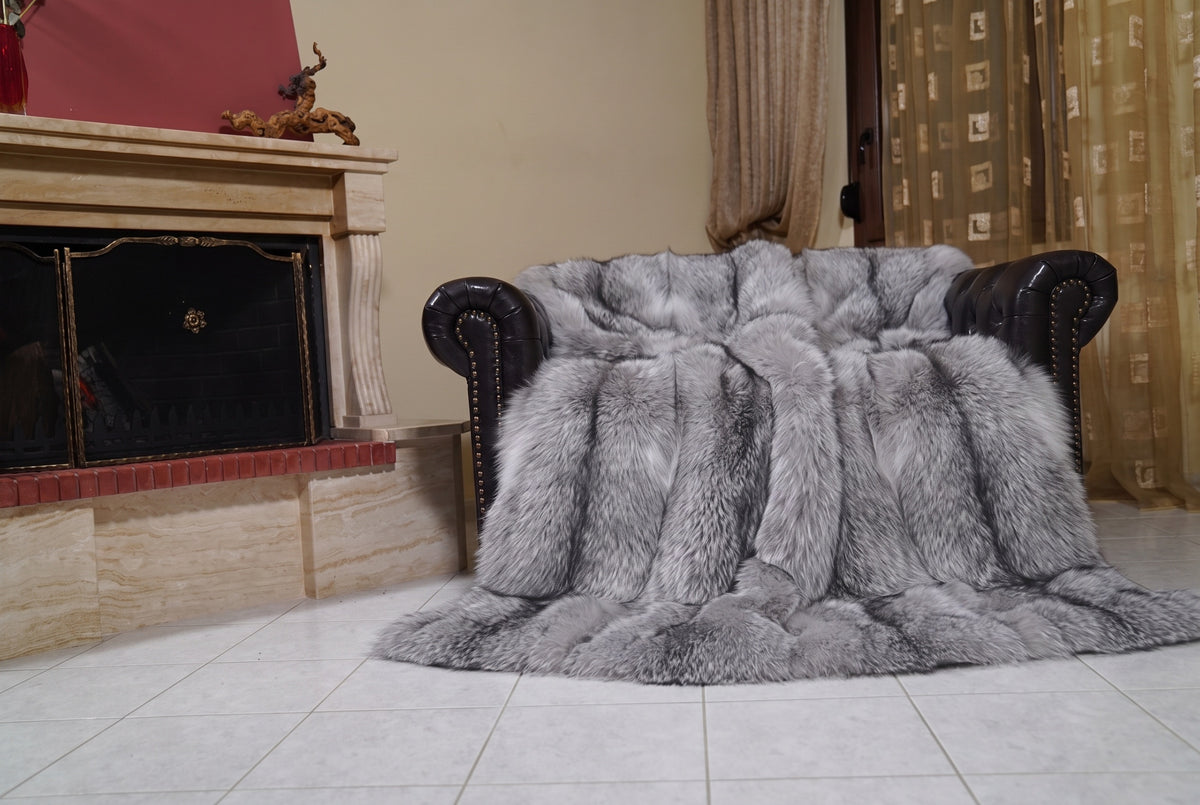 Fox Fur Blanket Throw Sapphire Frost — Full Skin Saga Furs draped over a dark brown leather Chesterfield armchair beside the fireplace — full view of the silver grey with white frosted tips and a charcoal line full skin fox pelts, TatiFurs