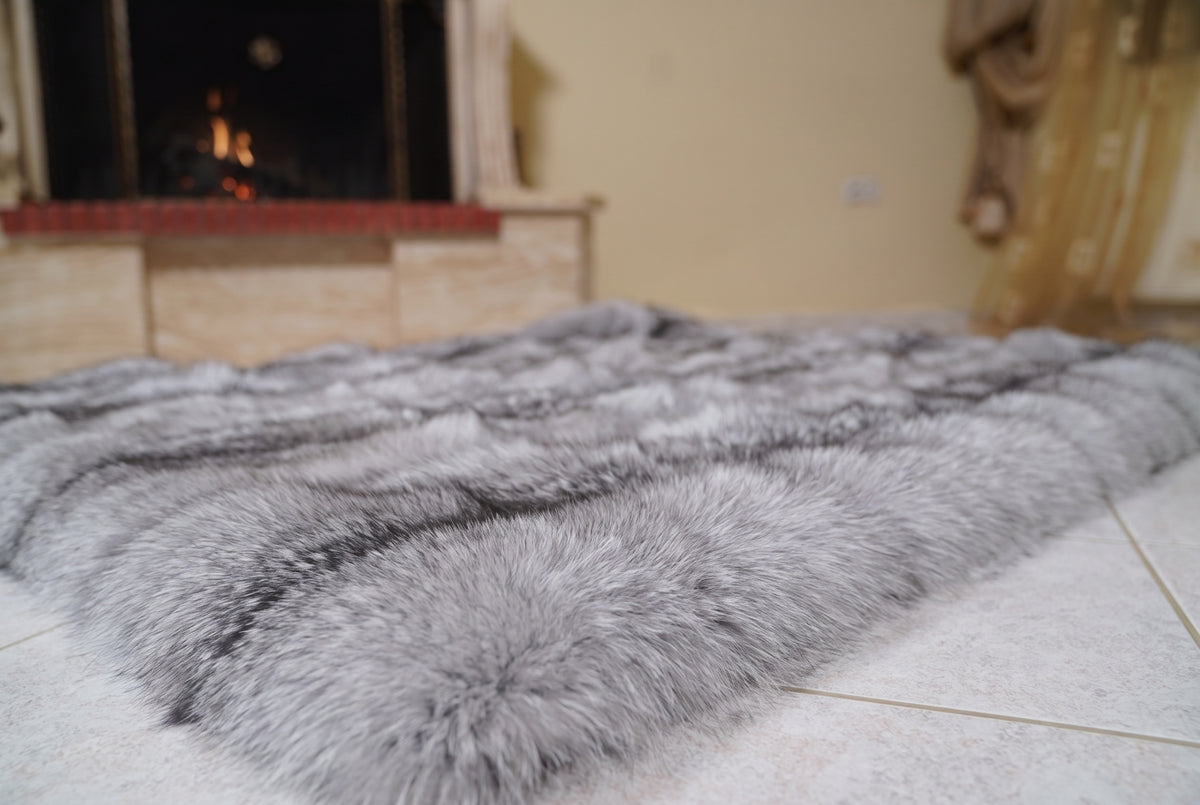 Fox Fur Blanket Throw Sapphire Frost — Full Skin Saga Furs laid flat on the floor in front of the lit fireplace, low angle showing the silver grey with white frosted tips and a charcoal line fox pelt rows, TatiFurs