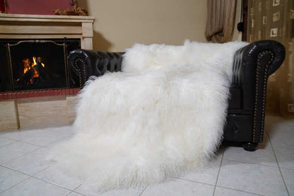 White Wool Sheepskin Mongolian Blanket Throw - Tatifurs draped over a black leather couch by a fireplace.