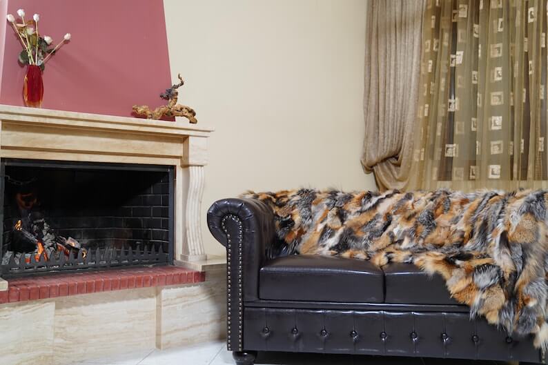 Fur Throw Blanket Natural Gross Fox – Luxury - Tatifurs displayed on a dark couch near a fireplace in a cozy living room.