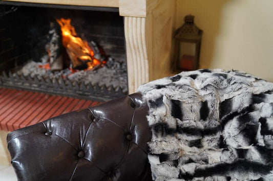 Rabbit Blanket Throw Chinchilla Rex - Tatifurs draped over a luxurious leather chair by a cozy fireplace.