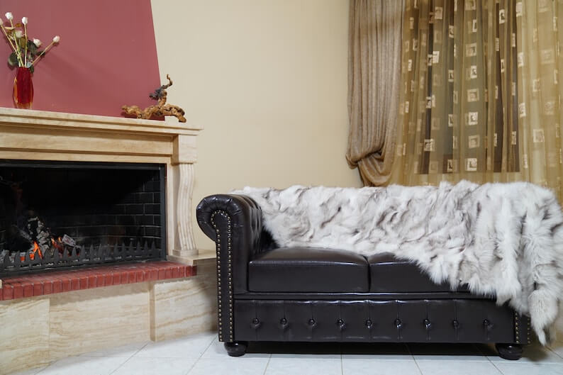 Fox Fur Blanket Throw Blue Natural - Tatifurs styled on a leather couch beside a fireplace.