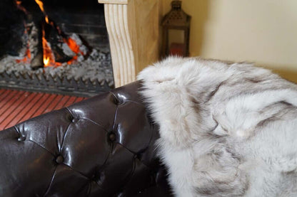 Fox Fur Blanket Throw Blue Natural - Tatifurs draped over a leather couch near a cozy fireplace.